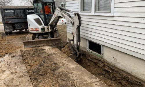 photo of a small white excavator digging along side the basement of a white house - an optional prep step to install Gorilla Wall Braces